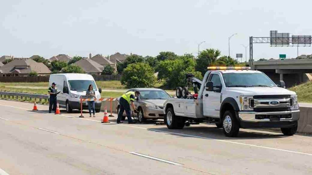 A professional roadside assistance team with a white tow truck and service van helping a stranded driver change a flat tire safely on a highway shoulder in Desoto, Texas.